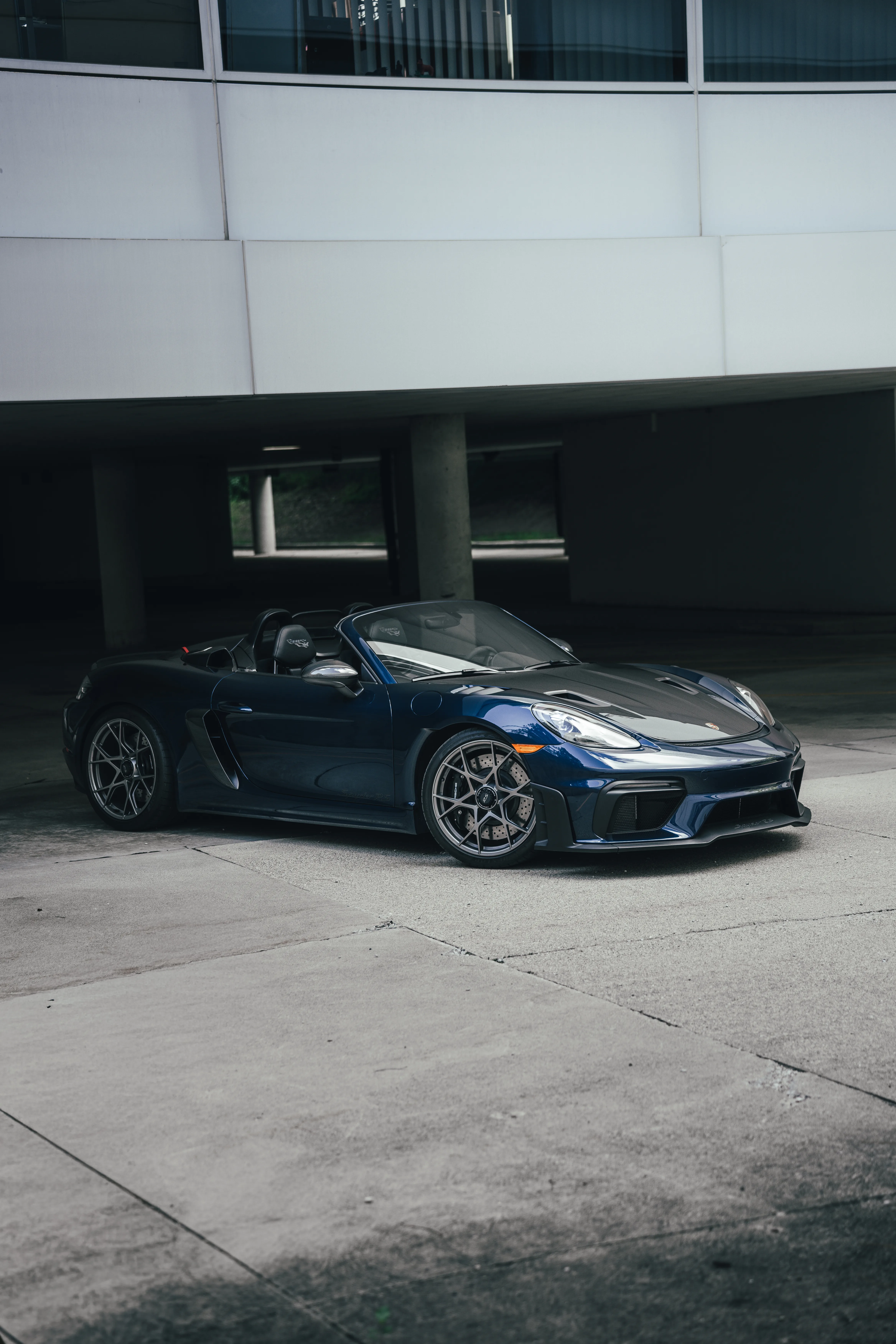 Front three-quarter view of a dark navy Porsche 718 Spyder RS parked beneath a parking garage overhang.