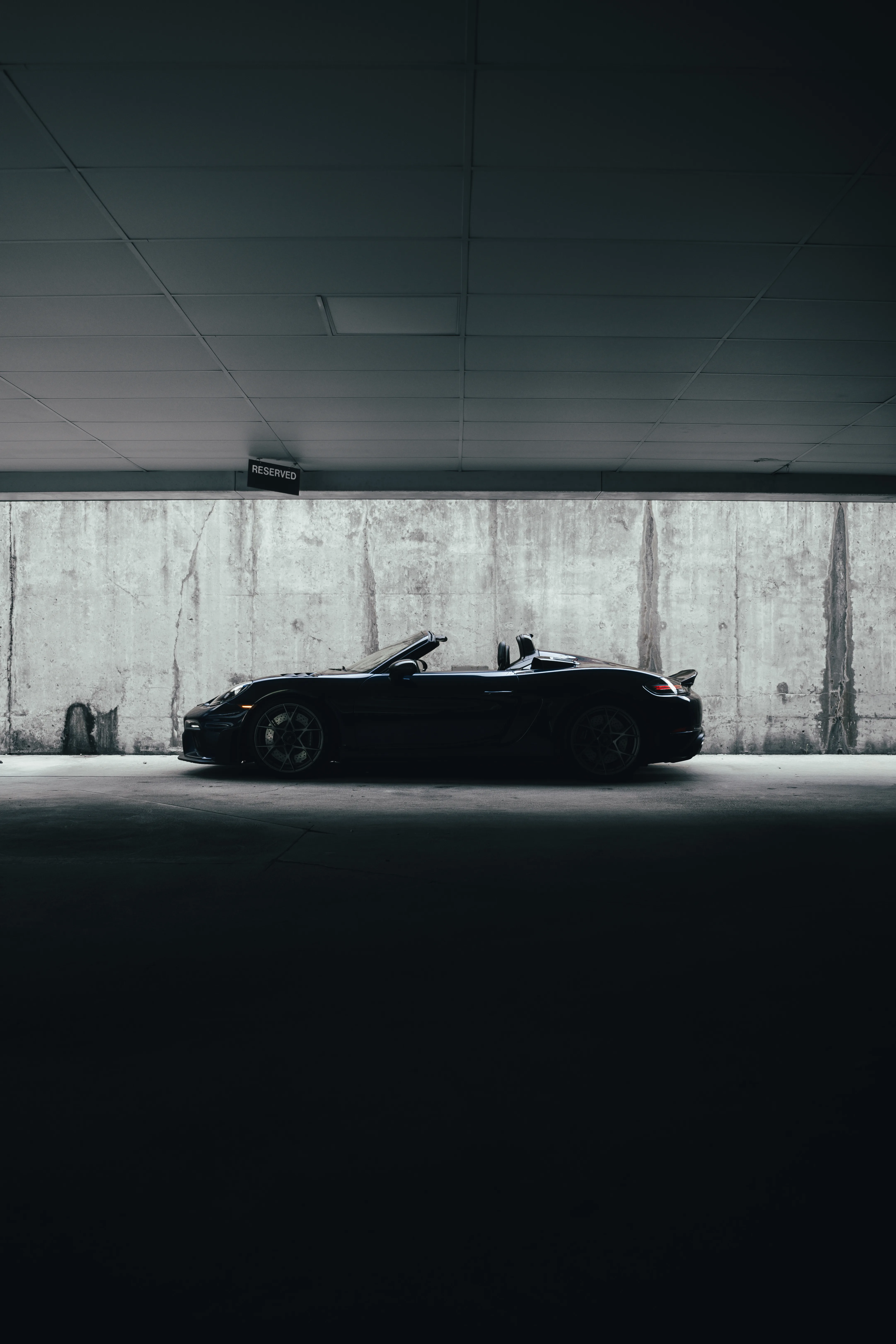 Side profile of a dark Porsche Spyder RS parked in a shadowed parking garage bay beneath a reserved parking sign.