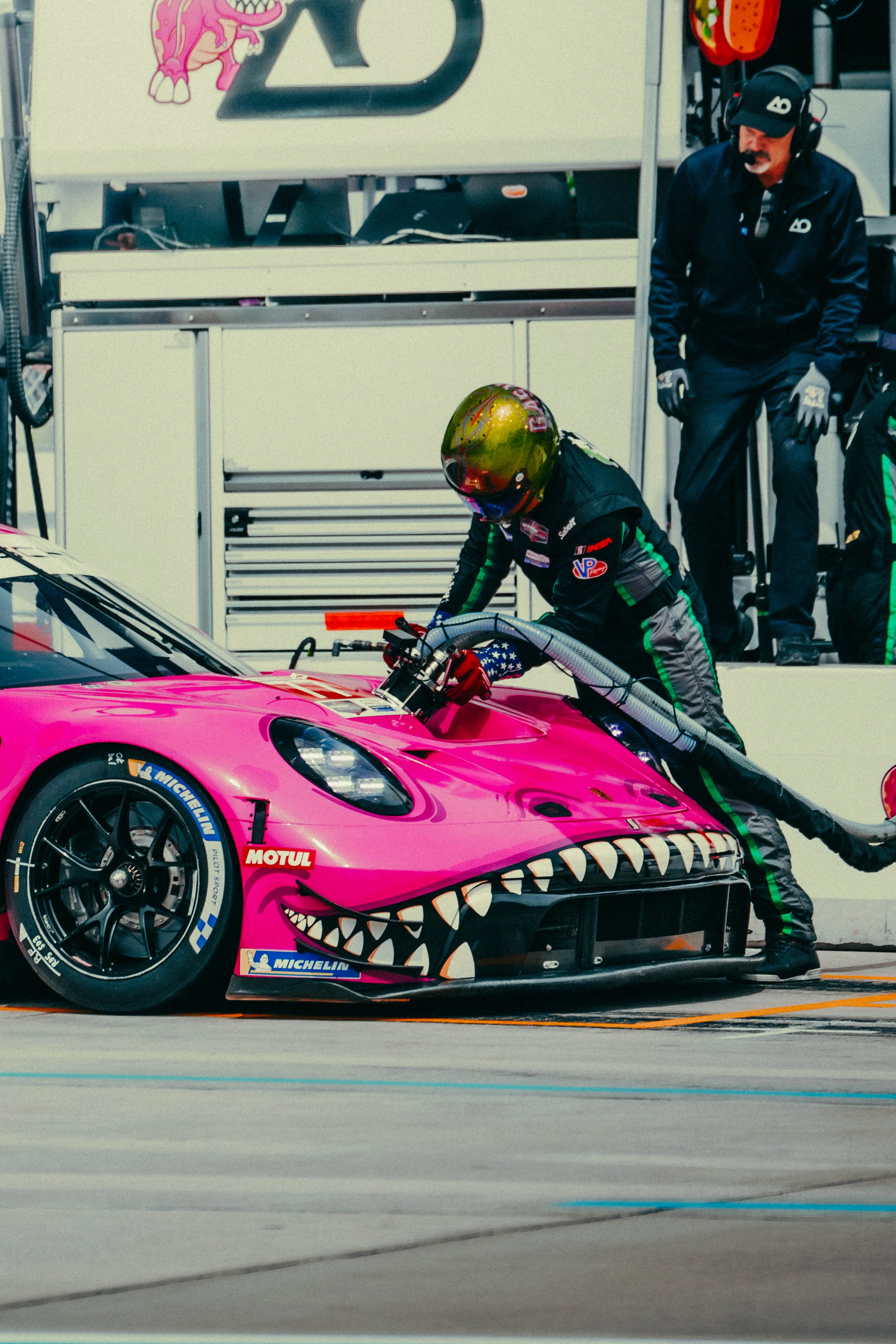 A pit crew member fuels a pink shark-mouth liveried GT race car in the pit lane, another crew member watching.