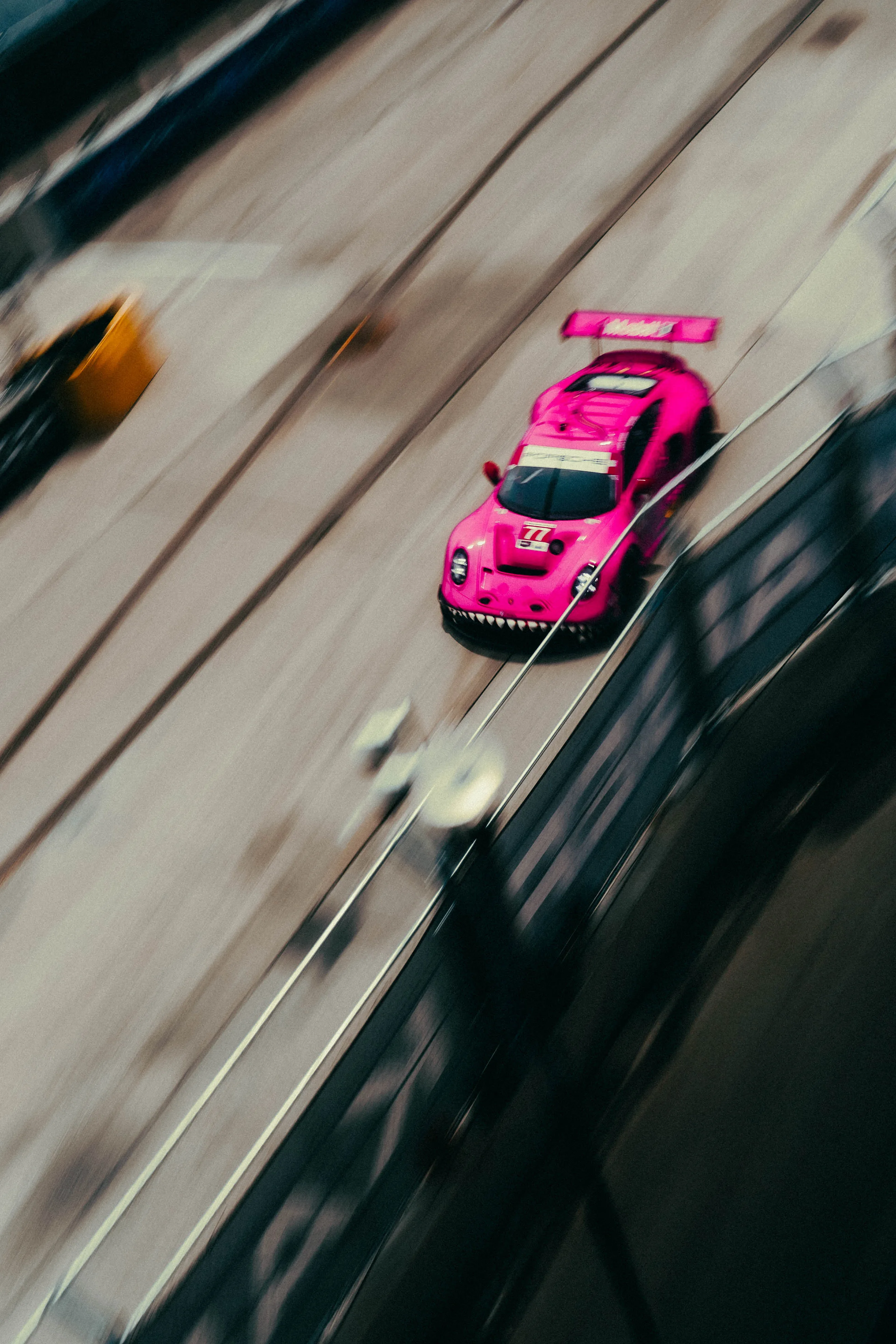 Panned action shot of a pink number 77 GT race car with a shark-mouth livery speeding down a street circuit.