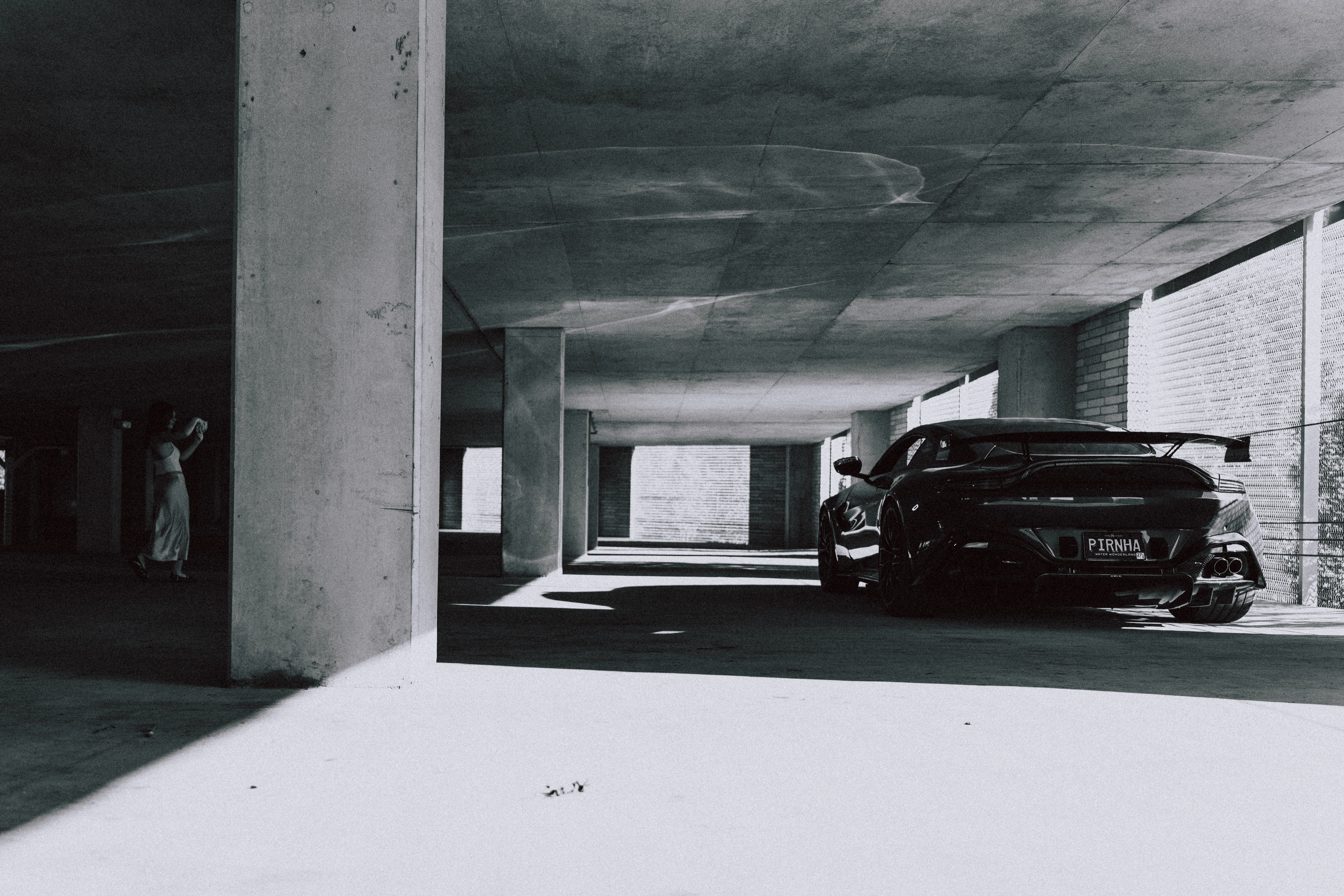Black and white shot of an Aston Martin's rear three-quarter in a parking garage, with a woman standing by a concrete pillar.
