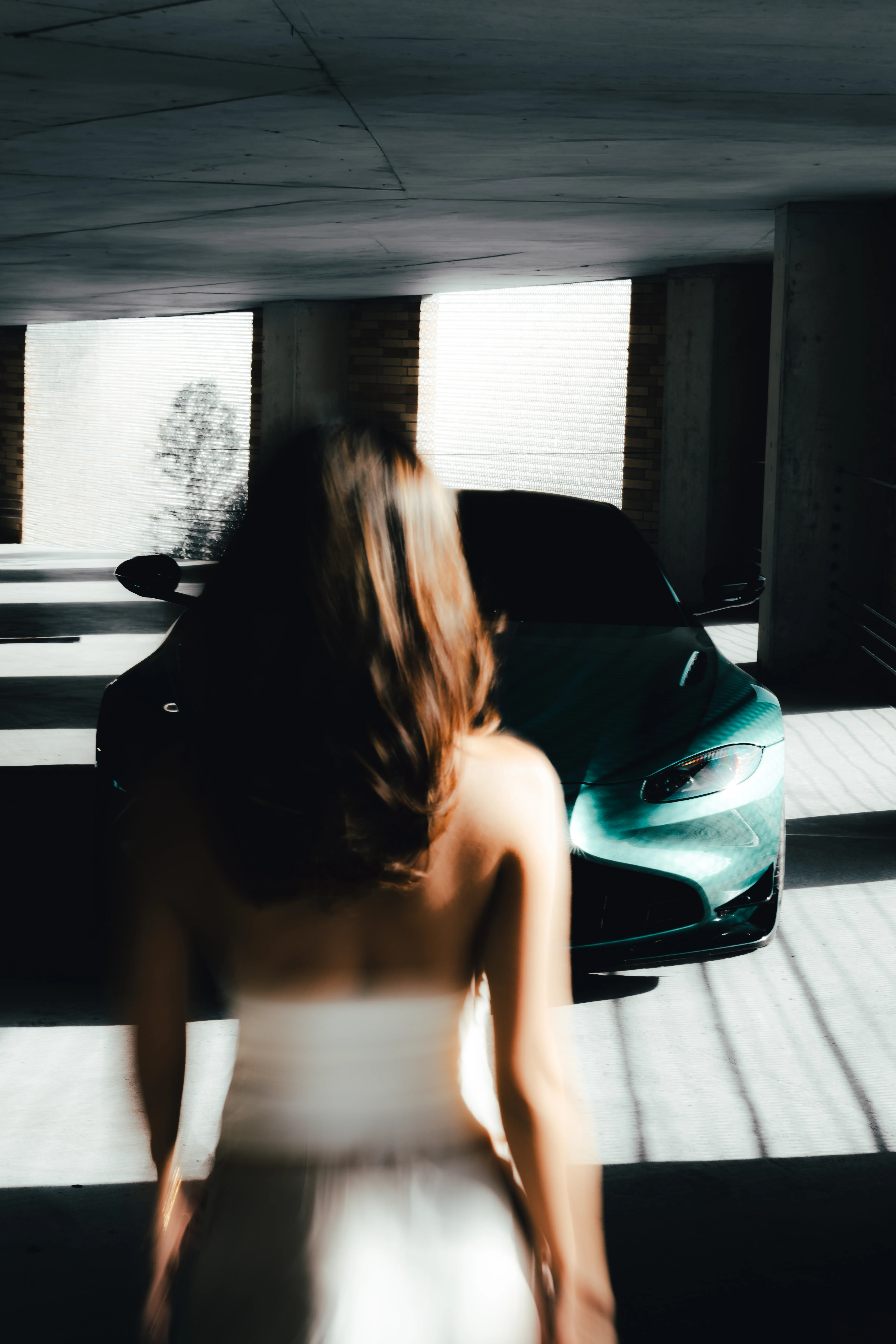 Front three-quarter view of a teal Aston Martin in a parking garage, with a woman in a white dress walking toward the camera.
