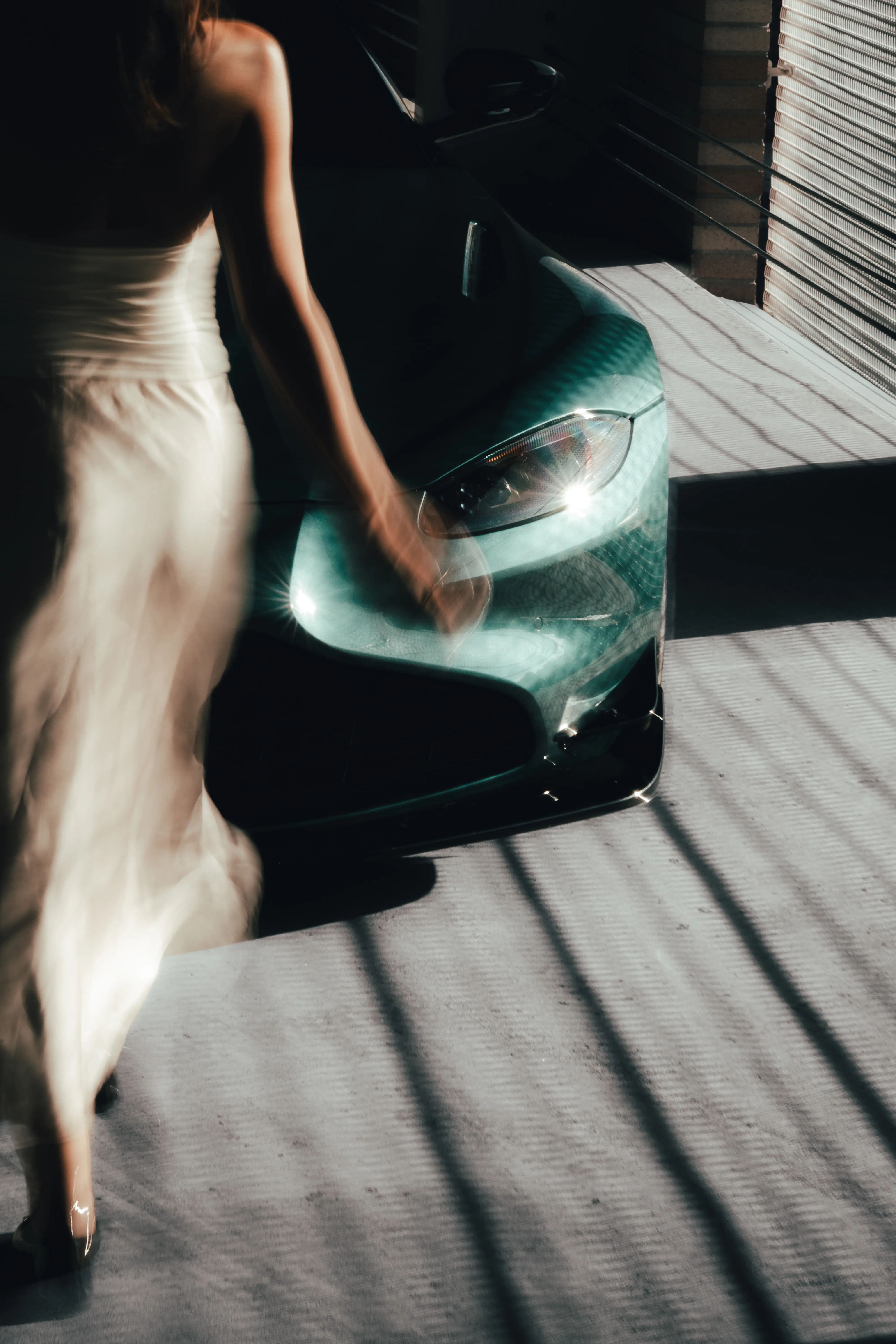 Close crop on an Aston Martin's headlight and hood, with a woman's hand and dress in the foreground.