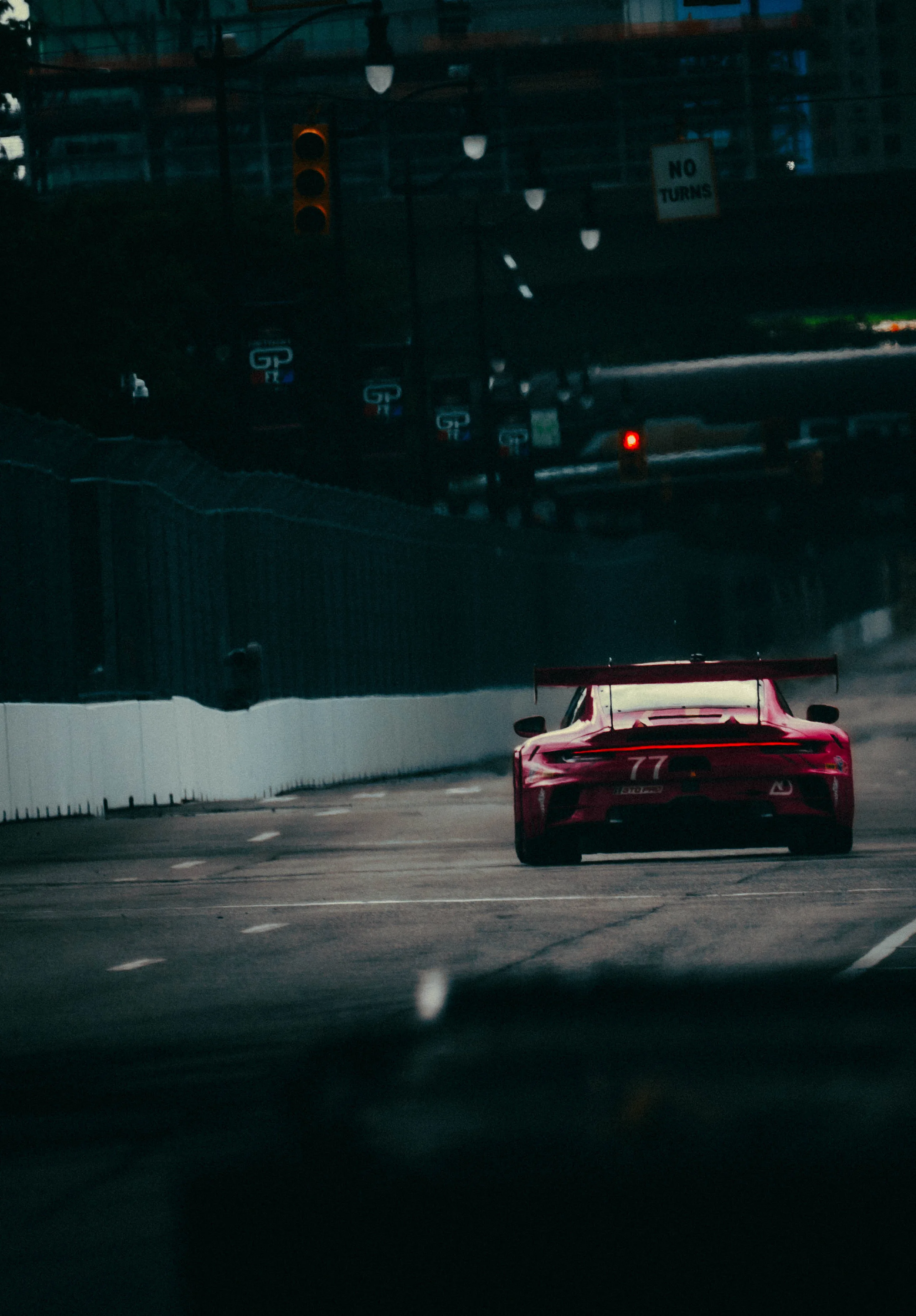Rear view of a red number 77 GT3 race car driving down a city street circuit at dusk, past a No Turns sign.