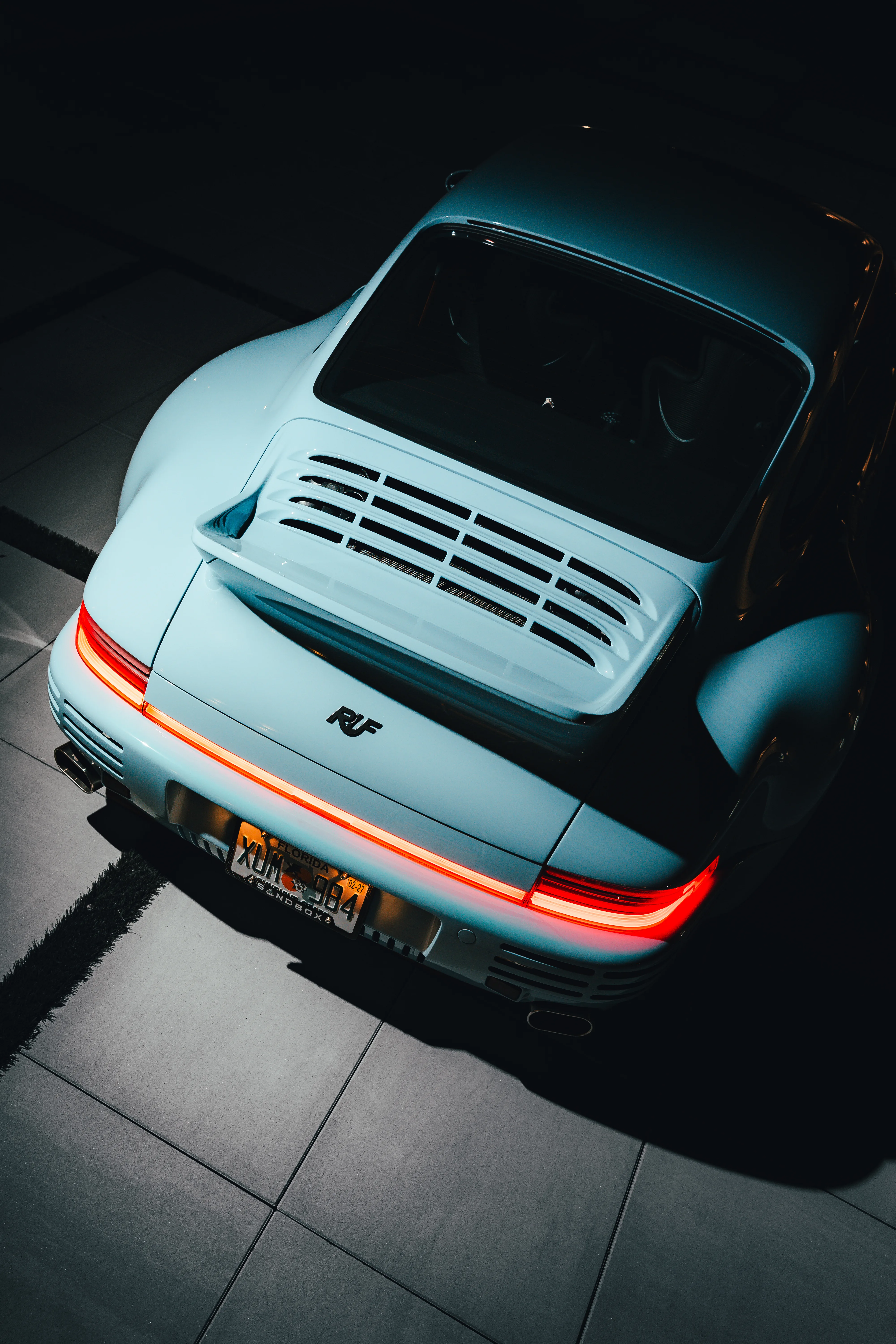 Rear detail of a light blue RUF Porsche at night, its full-width taillight lit red against the dark estate grounds.