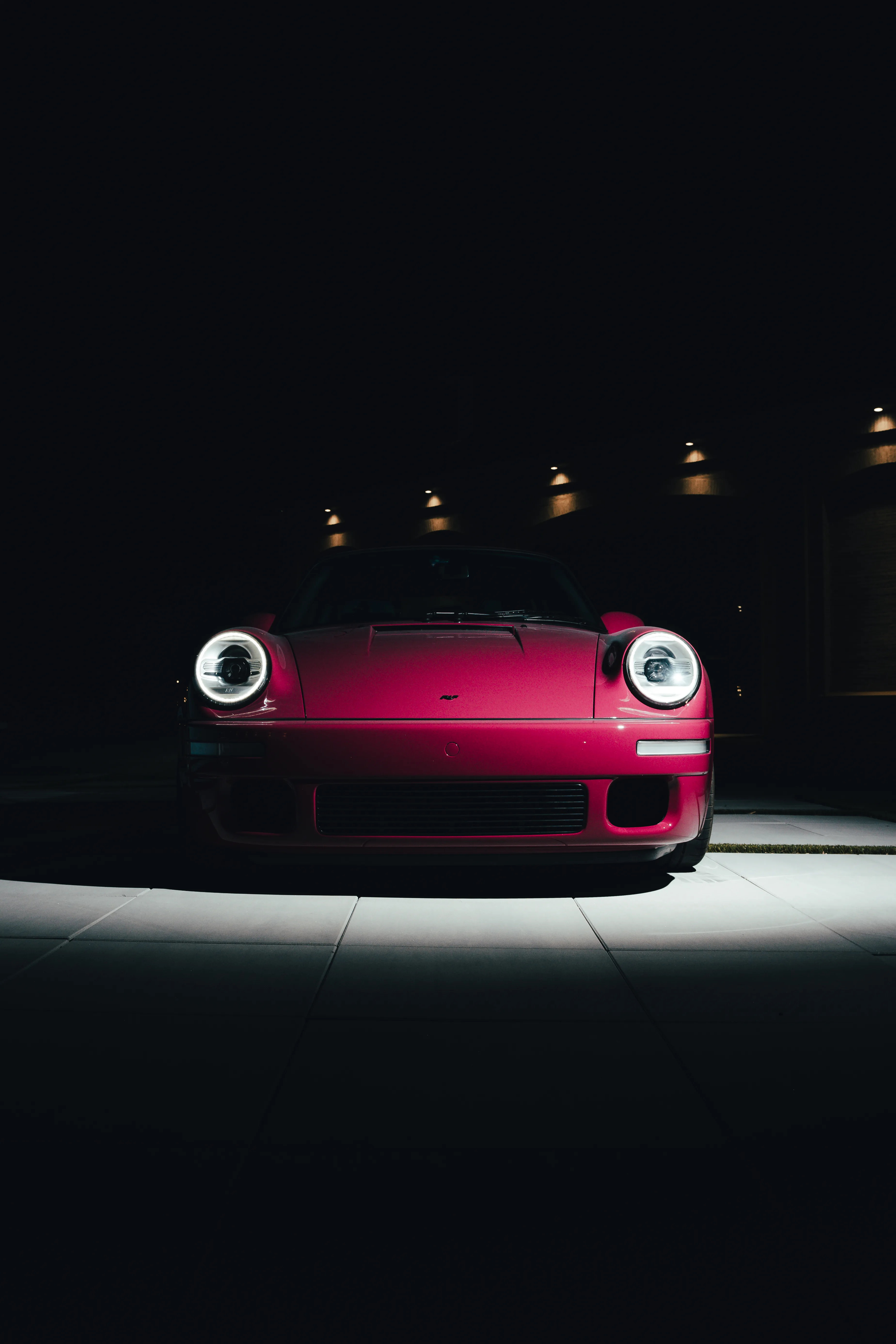 Front view of a magenta Porsche with round illuminated headlights, spotlit against a dark estate driveway at night.