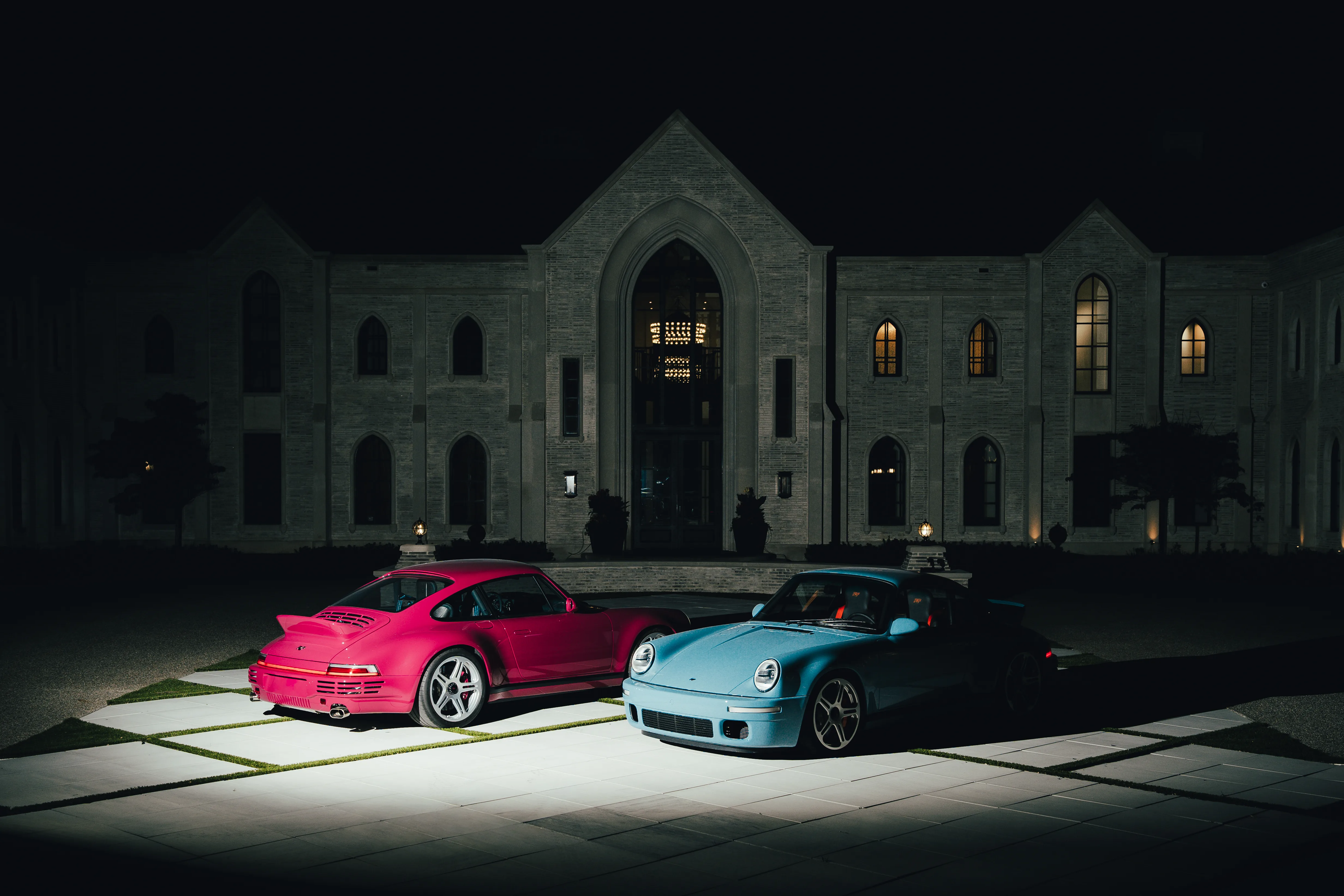 A magenta Porsche 959-style car and a light blue RUF parked at night in front of a large gothic-style stone estate.