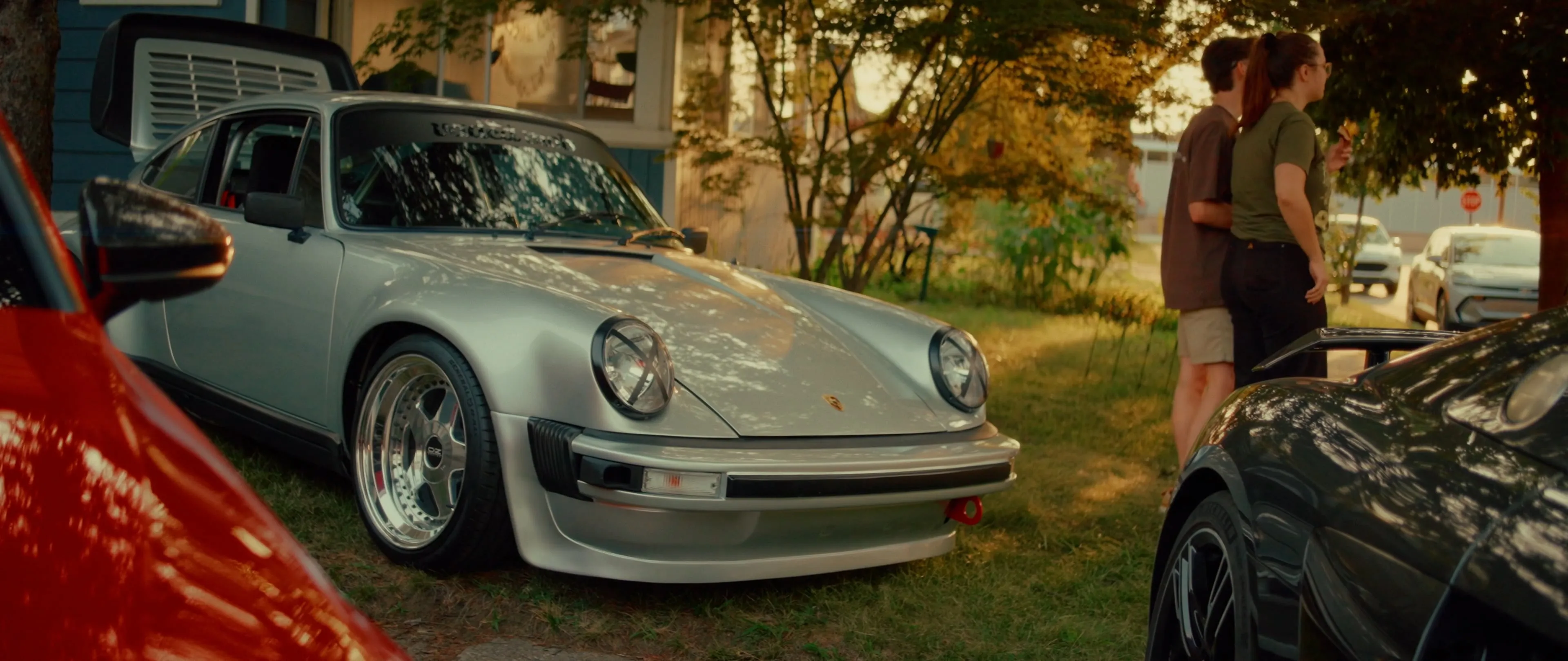 A silver Porsche 911 and a black car parked on a lawn in front of a house at golden hour, with two people standing nearby.