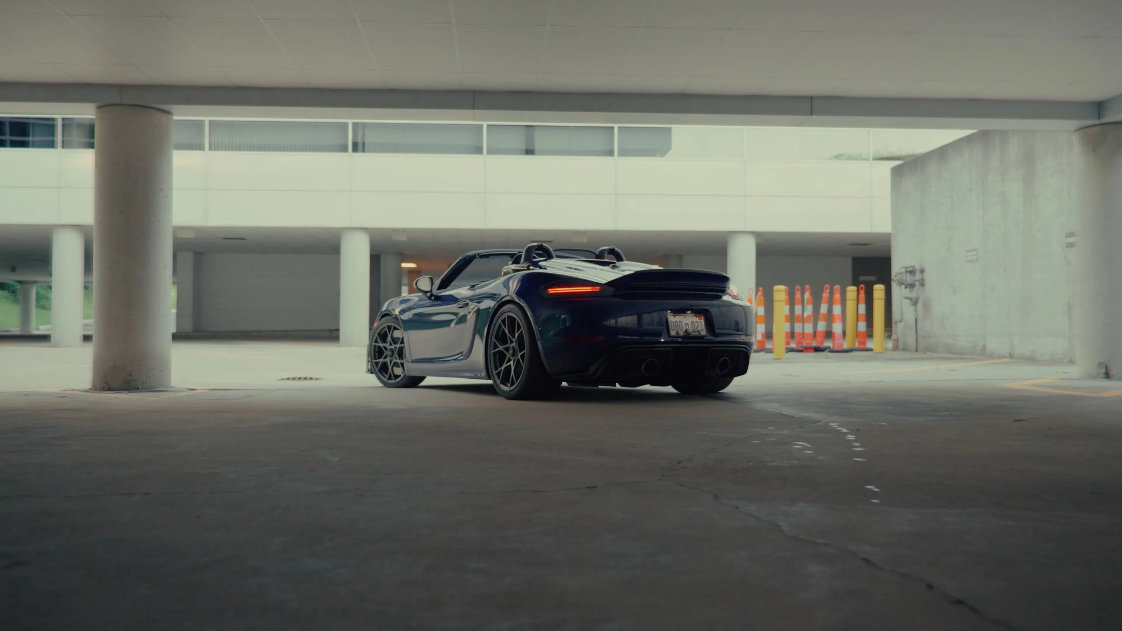 A dark blue Porsche 718 Spyder RS parked in a parking garage, rear three-quarter view, next to orange and yellow safety cones.