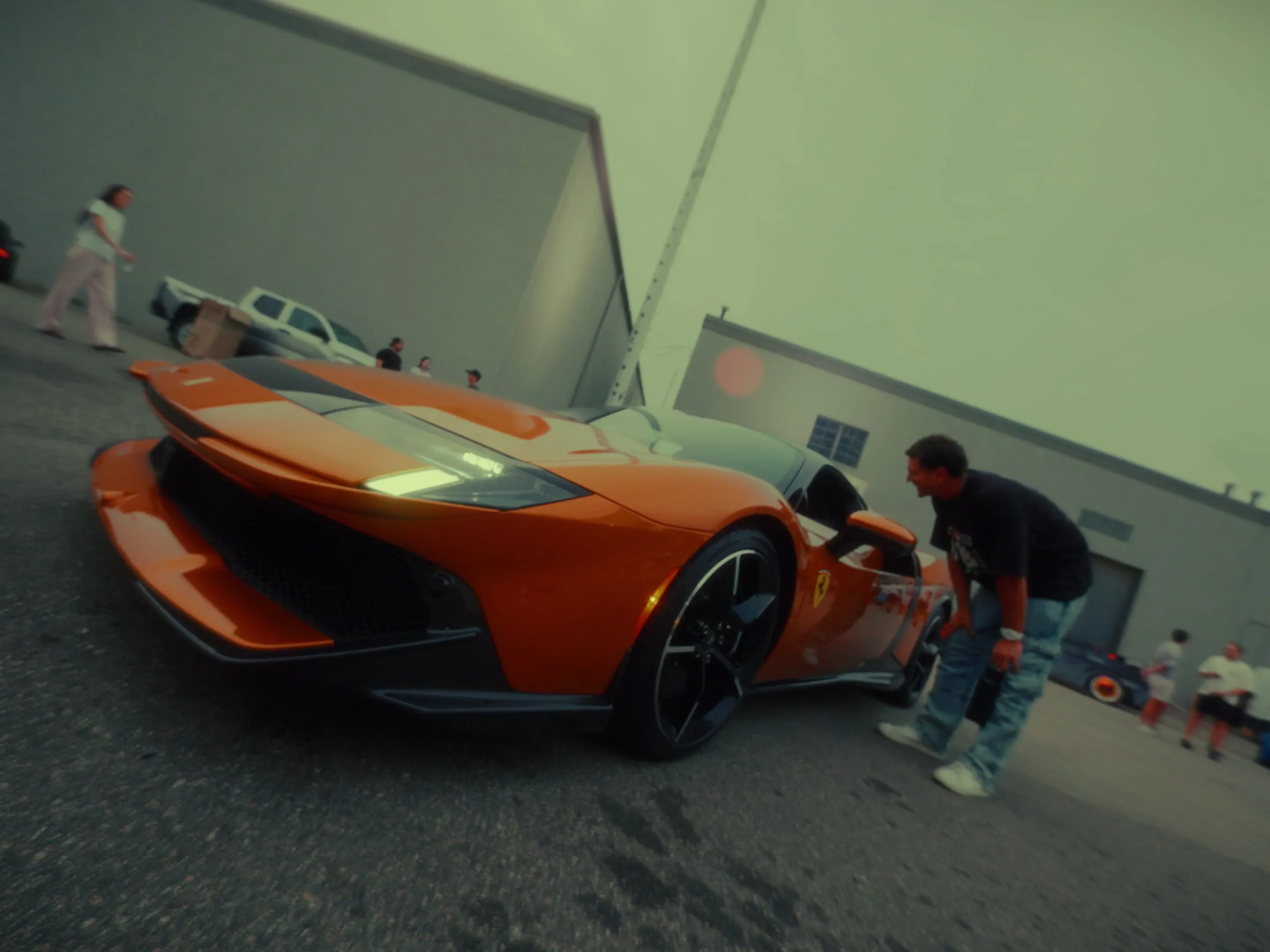 An orange Ferrari parked outside a warehouse at night, crew gathered around it.