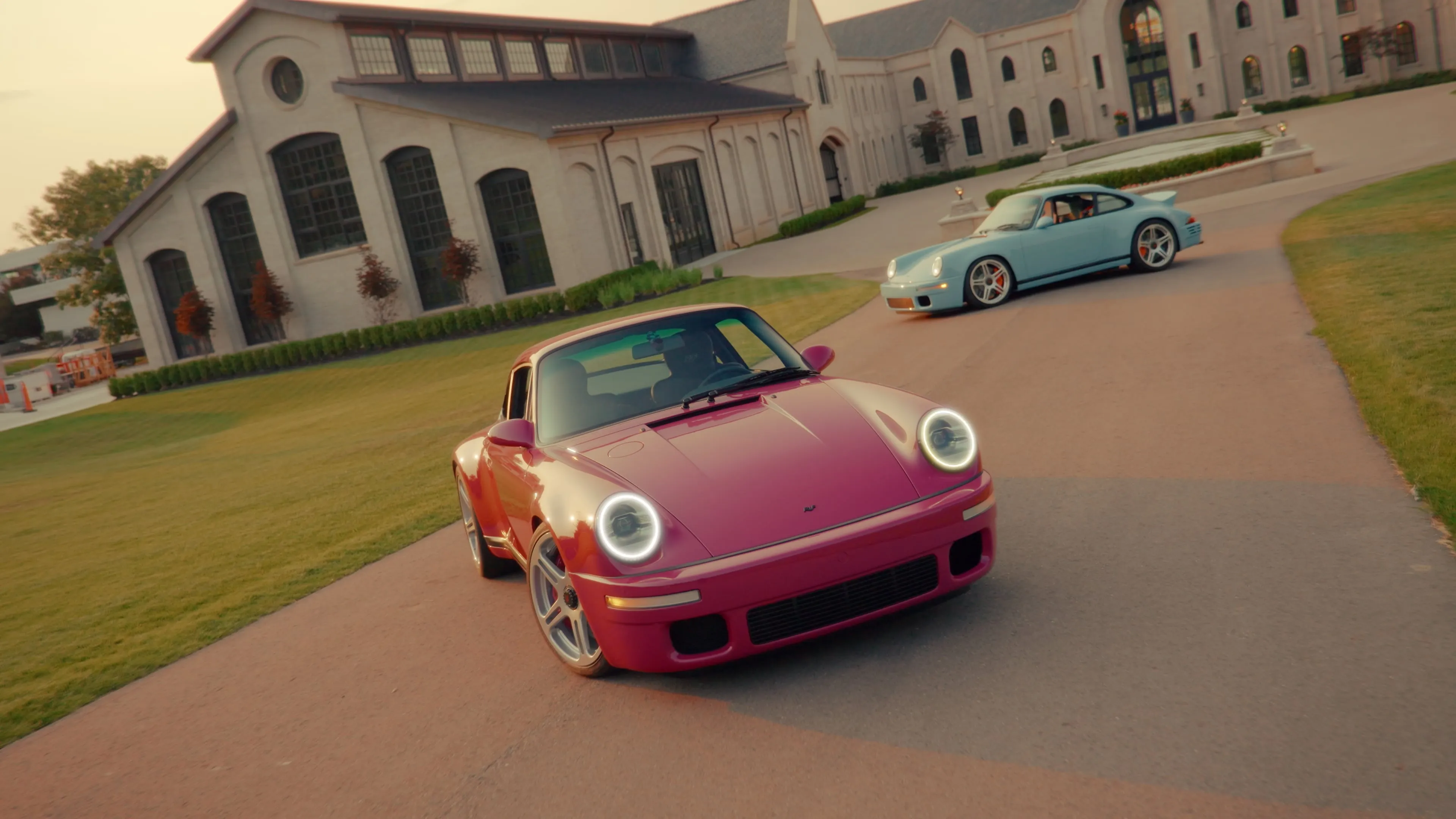 A pink RUF and a blue Porsche 911 parked on the driveway of a stone estate at golden hour.